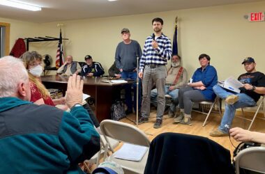 A speaker holding a microphone stands at the front of the town meeting and addresses the seated residents