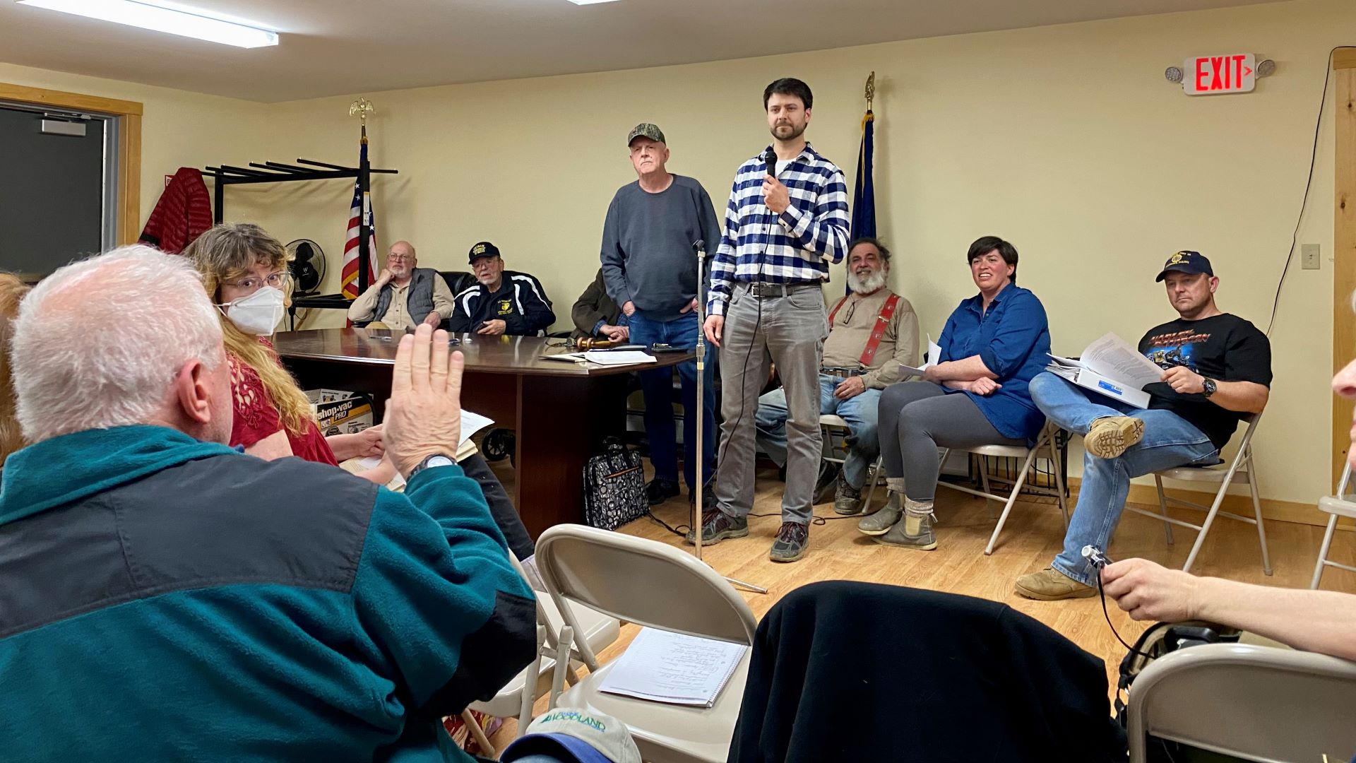 A speaker holding a microphone stands at the front of the town meeting and addresses the seated residents