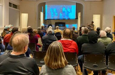 Dwayne Shaw speaks to a room full of seated individuals during a presentation with an image from the presentation being displayed on a projector screen.