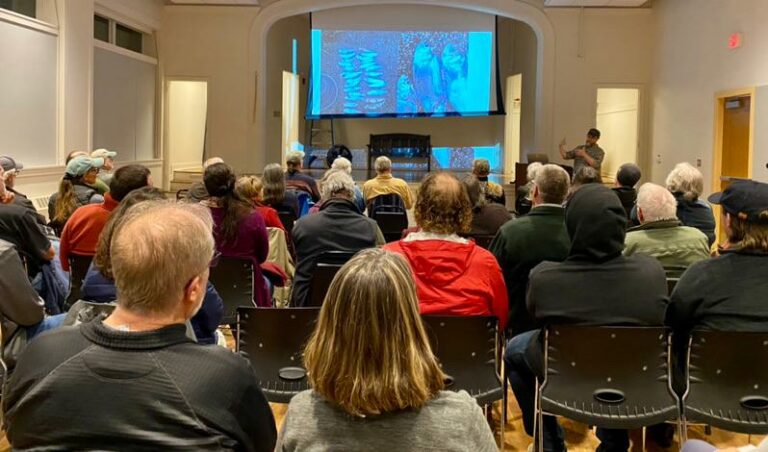 Dwayne Shaw speaks to a room full of seated individuals during a presentation with an image from the presentation being displayed on a projector screen.