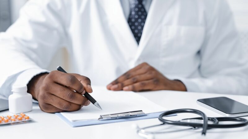 A Black man wearing a white doctor's coat fills out paperwork attached to a clipboard on a table.