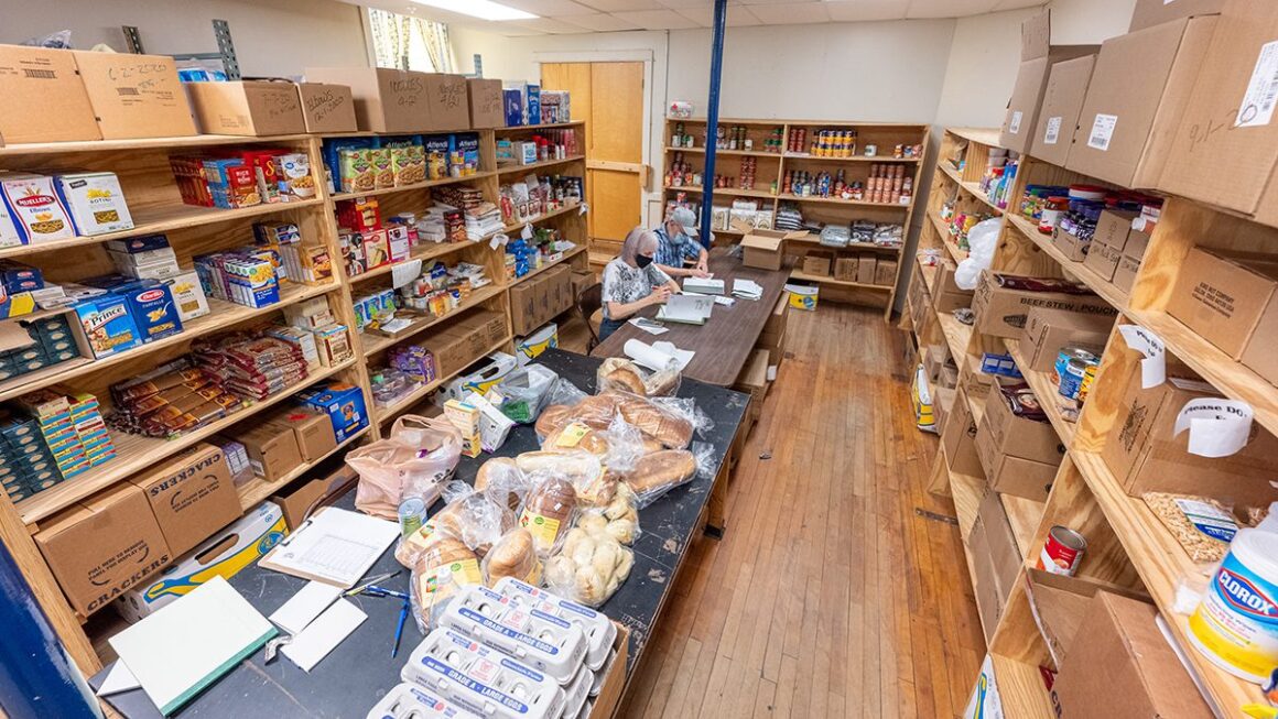 An overhead view of a room lined with stocked shelves inside the Come Spring Food Pantry with two individuals sitting at a table to assemble booklets.