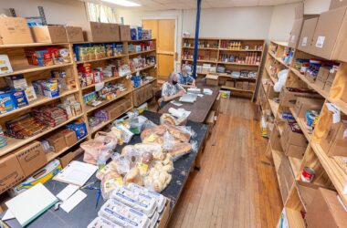An overhead view of a room lined with stocked shelves inside the Come Spring Food Pantry with two individuals sitting at a table to assemble booklets.