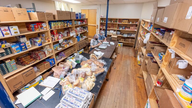 An overhead view of a room lined with stocked shelves inside the Come Spring Food Pantry with two individuals sitting at a table to assemble booklets.