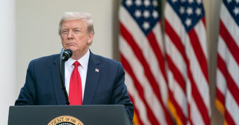 President Donald Trump stands at a podium during an event at the White House with a trio of U.S. flags behind him.