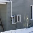 A heat pump seen attached to the side wall of a gray home with snow covering the ground.