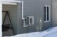 A heat pump seen attached to the side wall of a gray home with snow covering the ground.