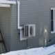 A heat pump seen attached to the side wall of a gray home with snow covering the ground.