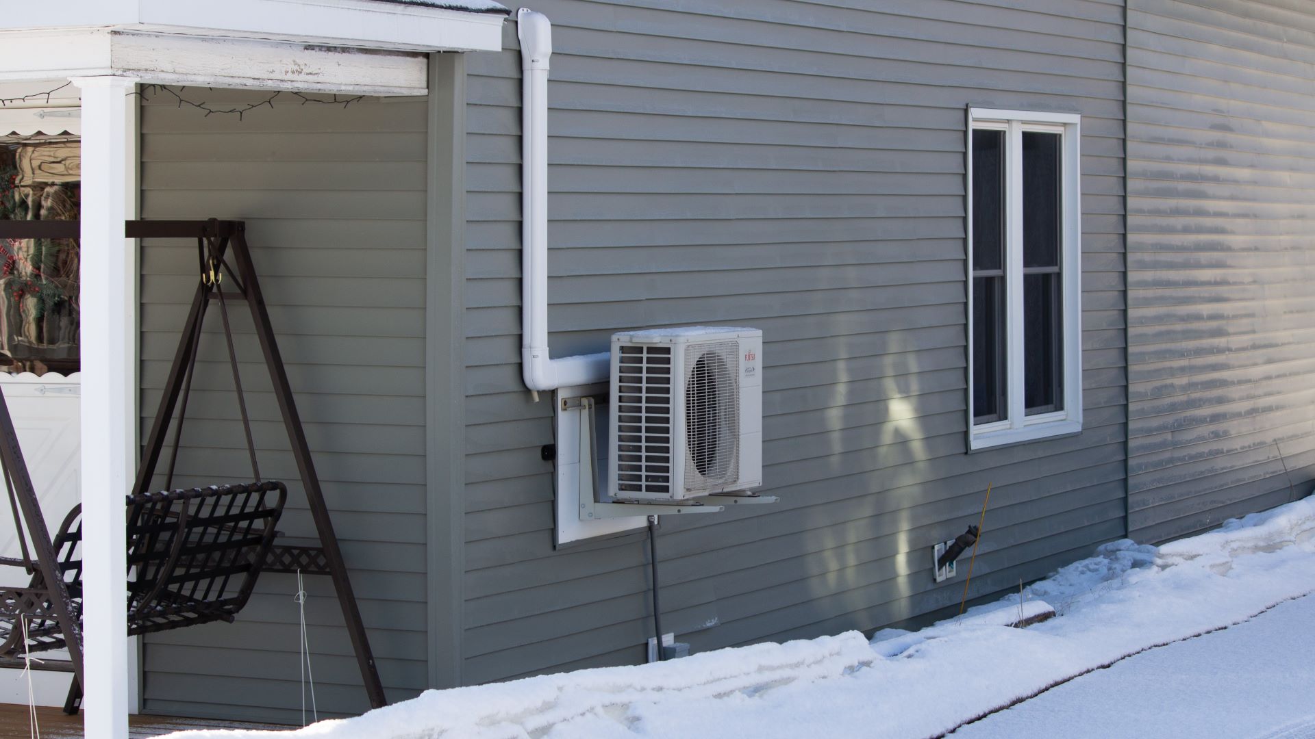 A heat pump seen attached to the side wall of a gray home with snow covering the ground.