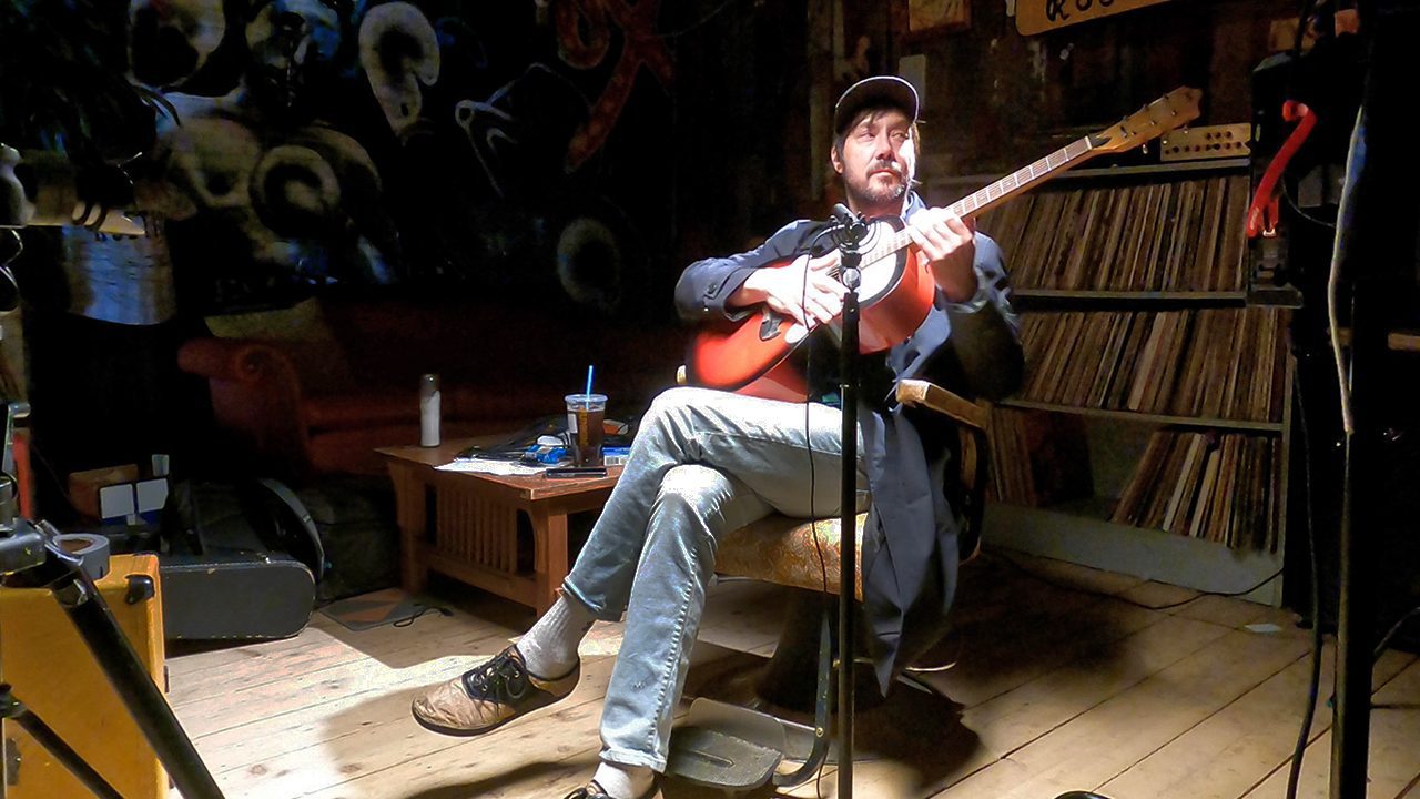 Dave Gutter plays a red guitar while sitting in a chair.