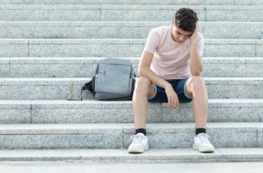 A young man sits on an outdoor staircase as he rests his head against his hand while looking down at the ground. His arm is resting on his knee.