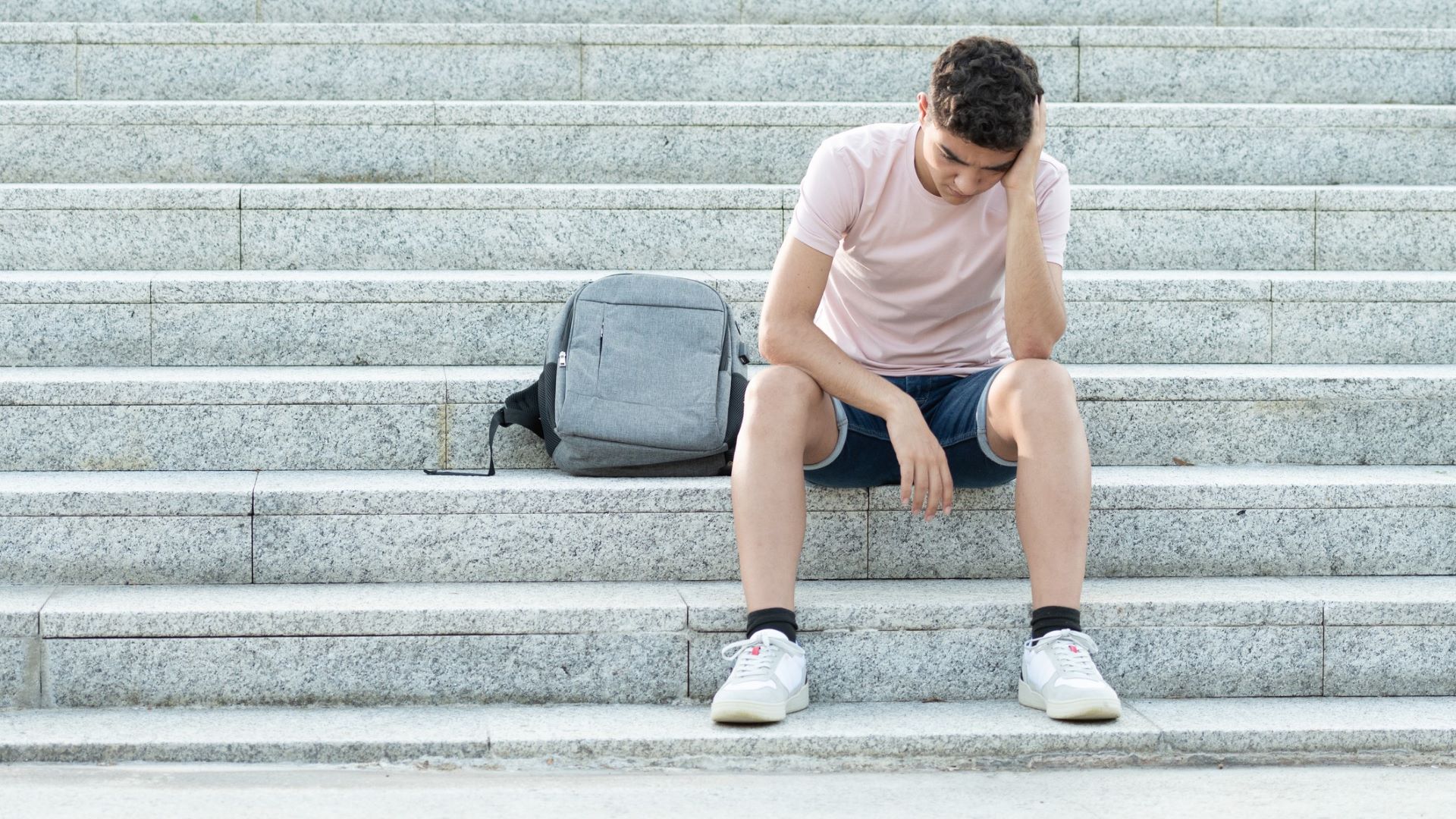 A young man sits on an outdoor staircase as he rests his head against his hand while looking down at the ground. His arm is resting on his knee.