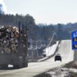 A logging truck passes a blue sign welcoming travelers to the Town of Baileyville and Village of Woodland.