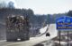 A logging truck passes a blue sign welcoming travelers to the Town of Baileyville and Village of Woodland.