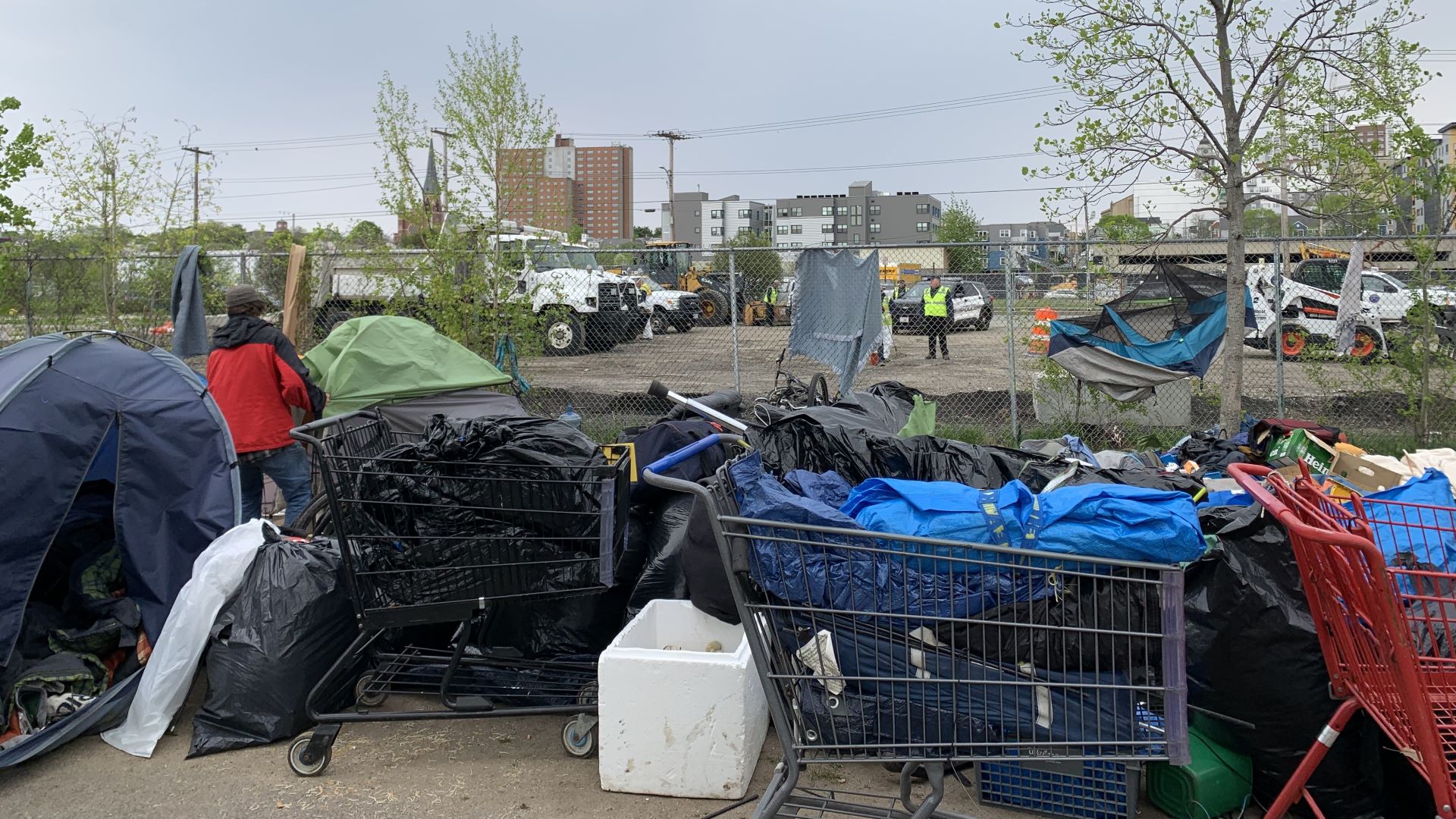 A man stands amid piled up bags of what was cleared from the Bayside encampment while beyond a chain-linked fence are idling bulldozers.
