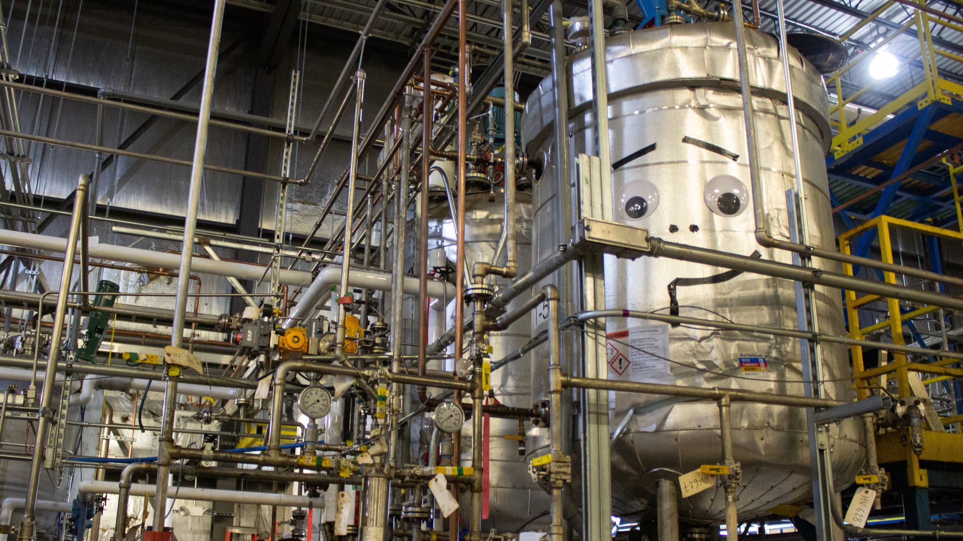 A tank with a makeshit face sits among many pipes.