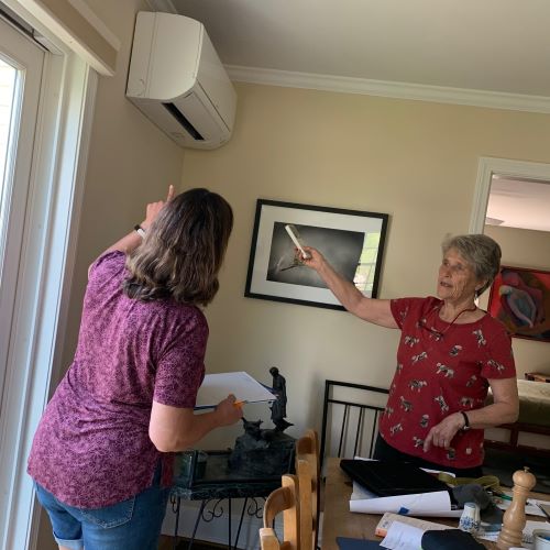Two women point to an installed heat pump inside a home.