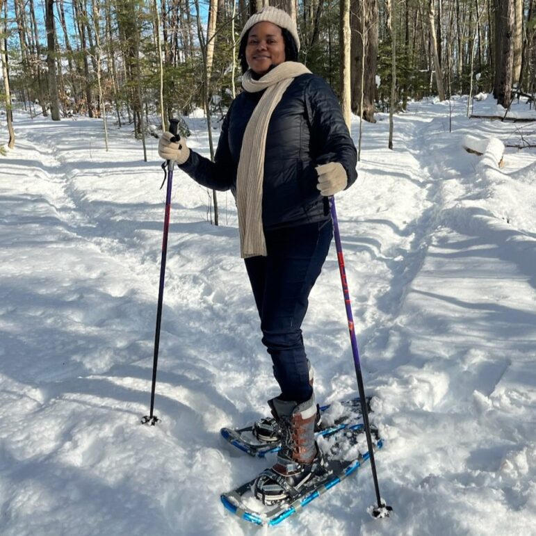 A woman poses for a photo while snowshoeing and surrounded by trees. 