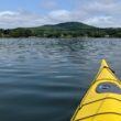 The front portion of a yellow kayak is seen on the open water with a mountain in the background.