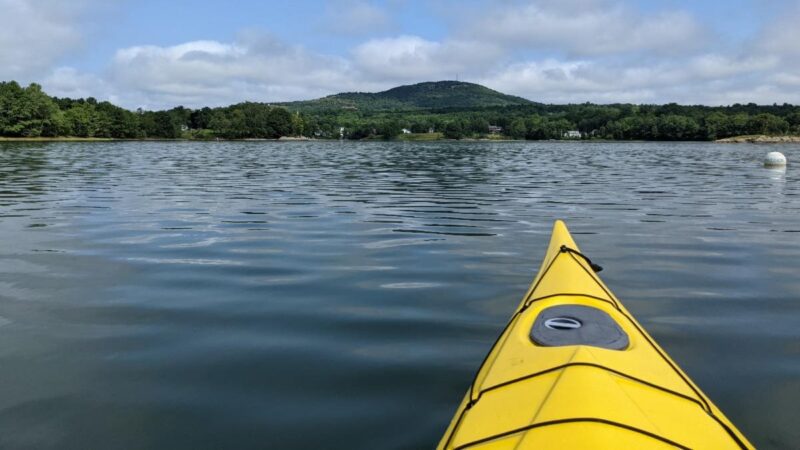 The front portion of a yellow kayak is seen on the open water with a mountain in the background.