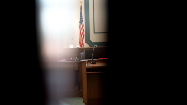 A small portion of an empty court room is visible through a cracked open doorway.