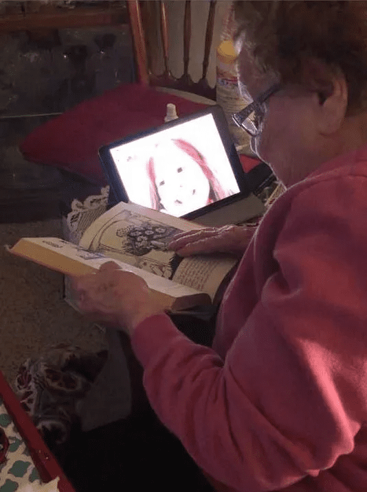 A woman reads a book while her young granddaughter's face fills her laptop screen.