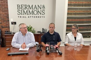 Michael Bigos, David Cole and Kimberly Cole sit at a conference table in a law firm's office during a press conference.