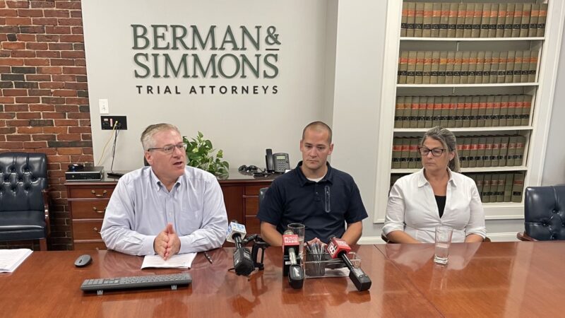 Michael Bigos, David Cole and Kimberly Cole sit at a conference table in a law firm's office during a press conference.