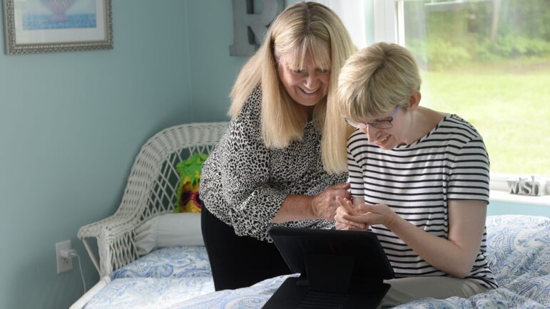Debbie Dionne and Kate Riordan smile while looking at an ipad and sitting on a bed.