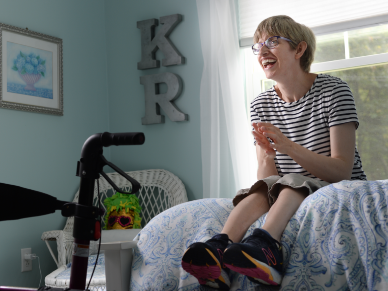 Kate Riordan smiles widely while sitting on the edge of her bed.