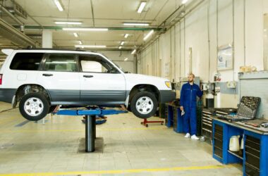 A car mechanic stands near a white car that is raised on a platform in an auto repair shop.