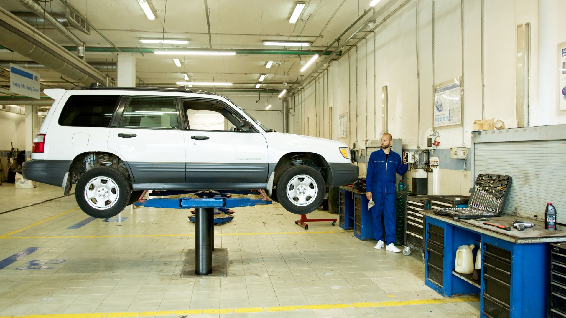 A car mechanic stands near a white car that is raised on a platform in an auto repair shop.