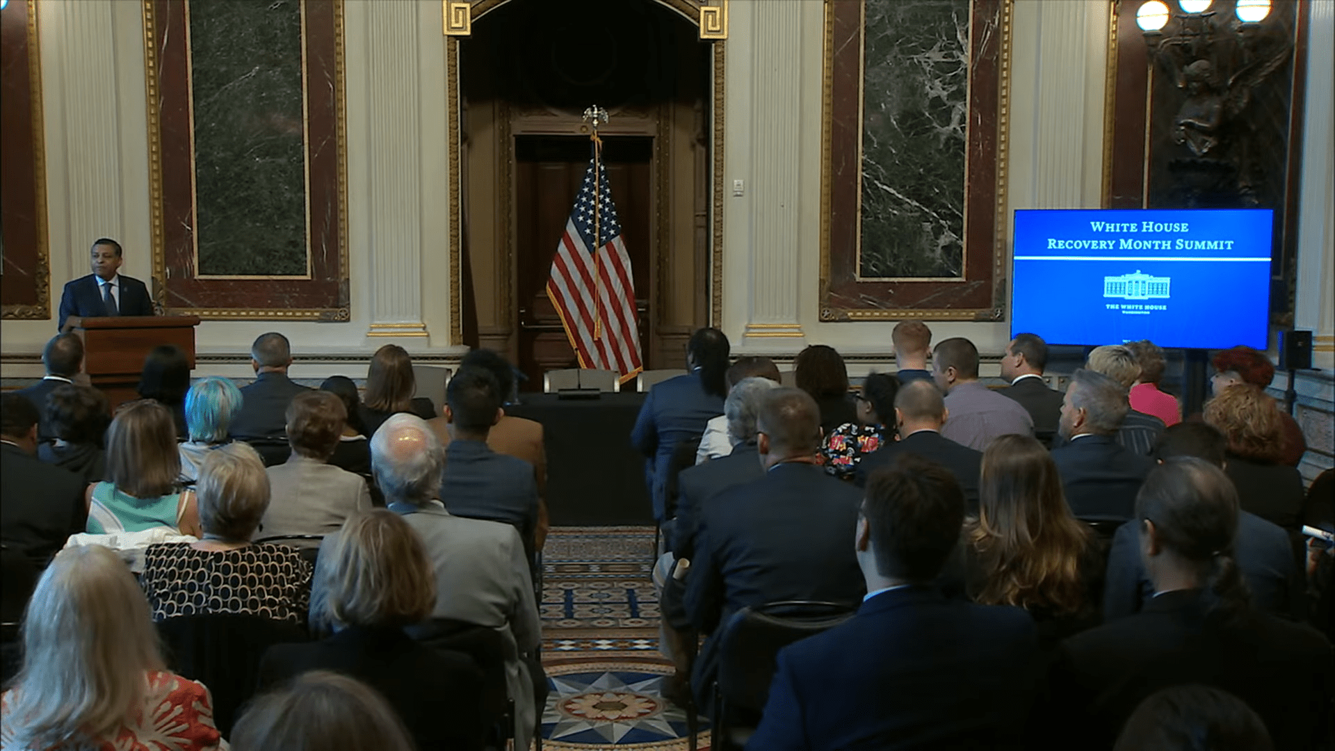 Dr. Rahul Gupta stands at a podium located in the front of a crowded room.