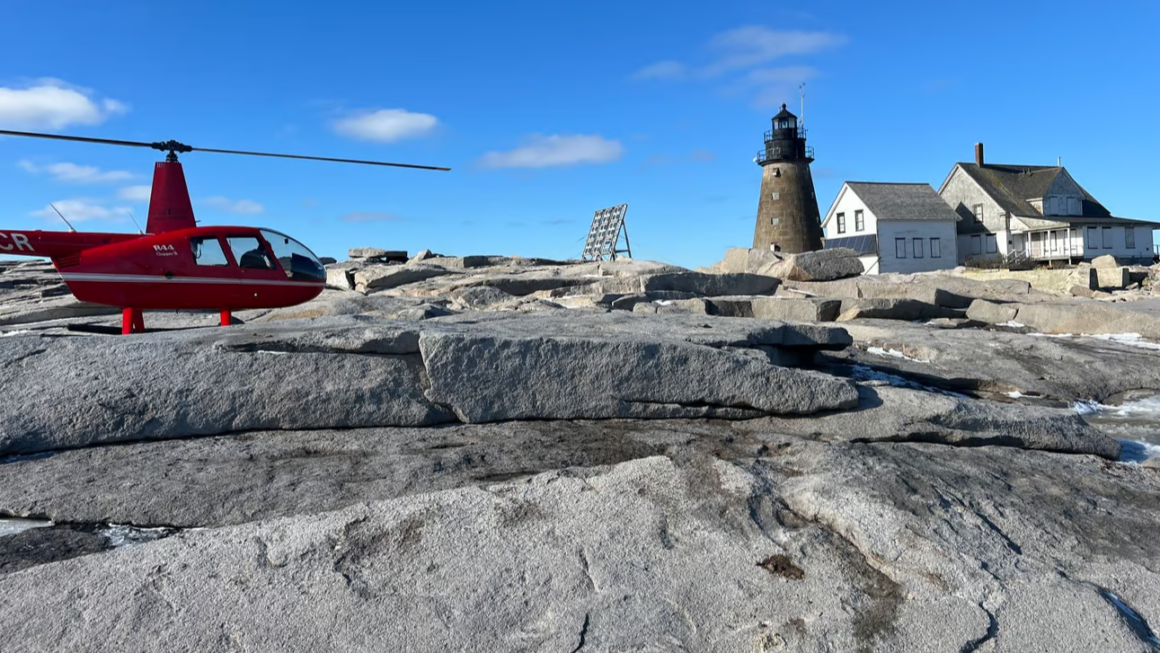 A red helicopter on the rocks by a lighthouse.