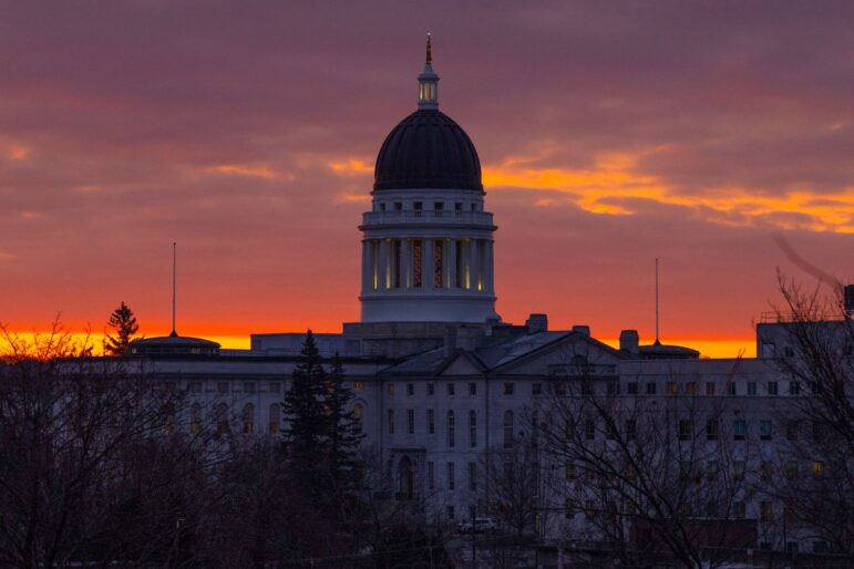 The exterior of the Maine State House seen against a slightly orange sky