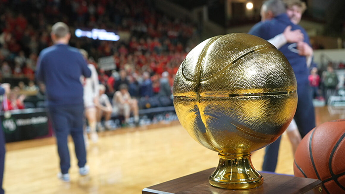 The gold basketball trophy sits on a table while players and coaches celebrate in the distance.