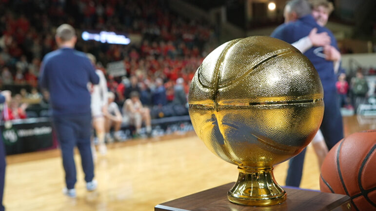 The gold basketball trophy sits on a table while players and coaches celebrate in the distance.