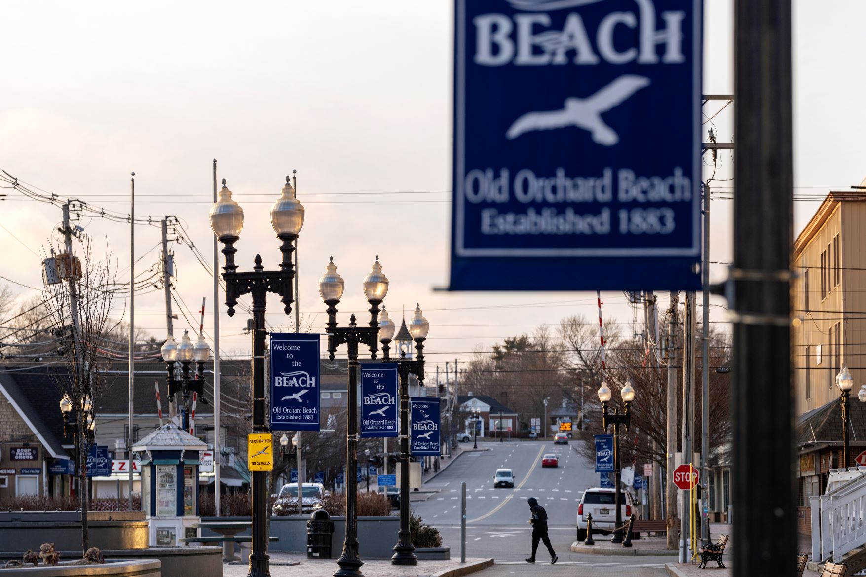 A man crosses a street in Old Orchard Beach. Hanging from several lamp posts are blue signs promoting the town of Old Orchard Beach.