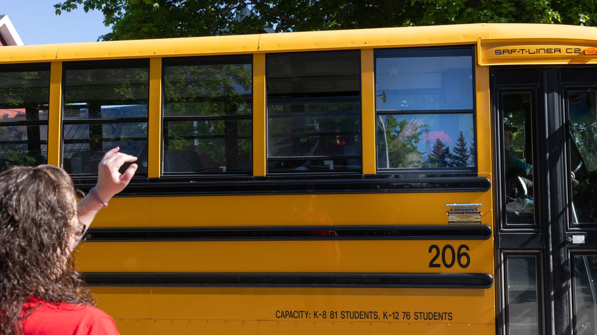 A mother waves goodbye to her daughter who is looking through the window of a school bus.