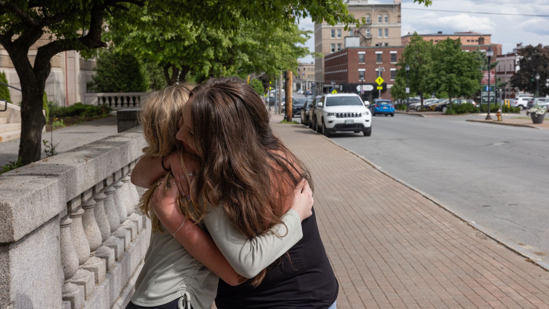 A mother and daughter embrace in a hug while standing on a sidewalk.