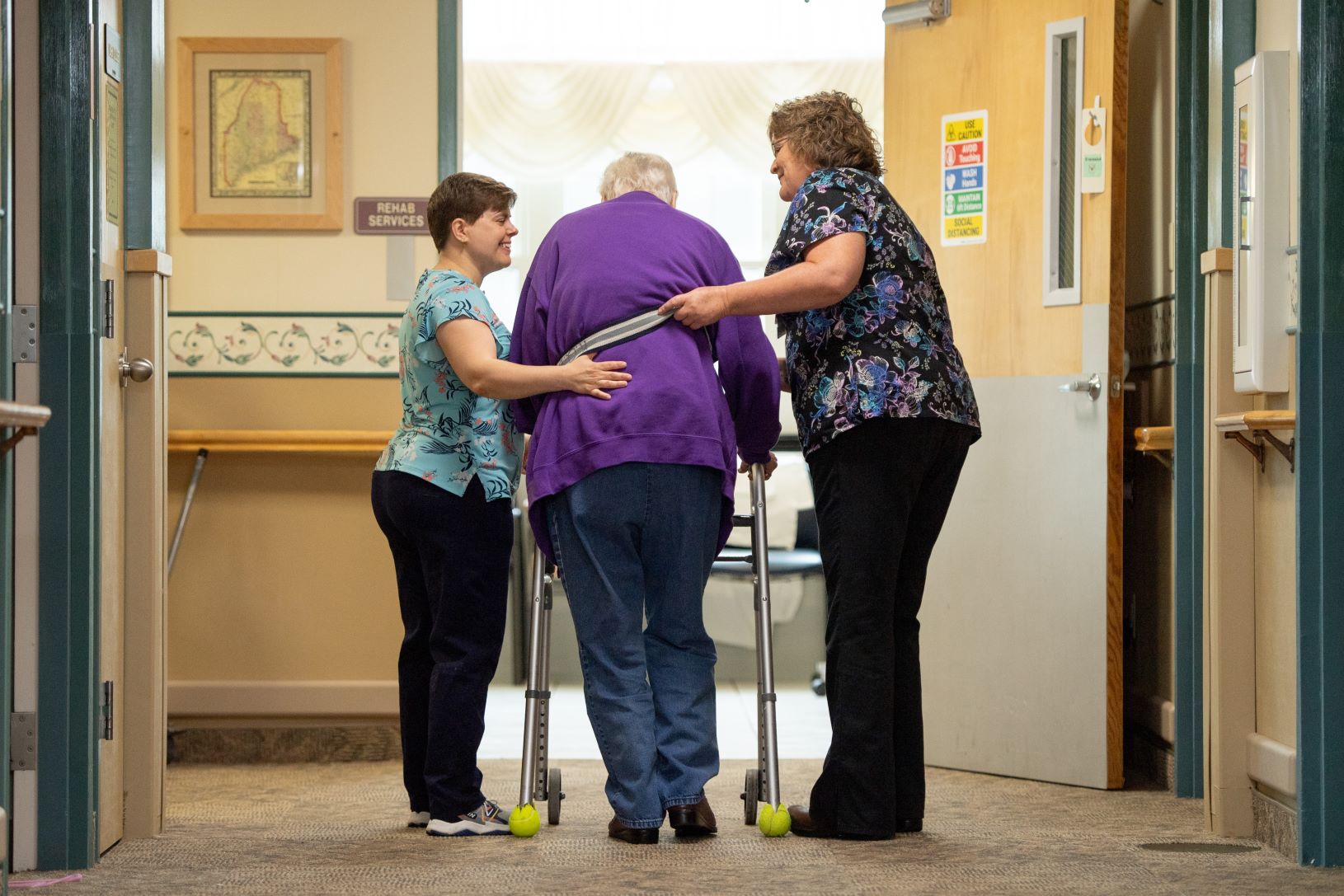 Two nurses assist a patient as the patient walks with the use of a walker.