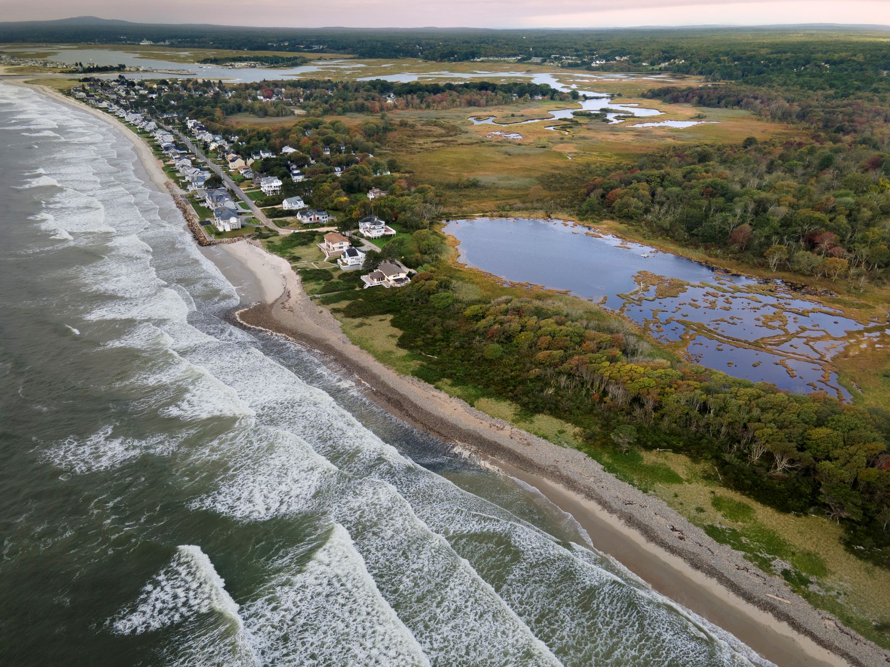 An aerial image showing waves crashing against the beach with houses along the beach in the near distance.