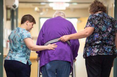 Two nurses help a patient walk with a walker.