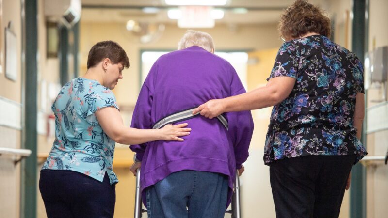 Two nurses help a patient walk with a walker.