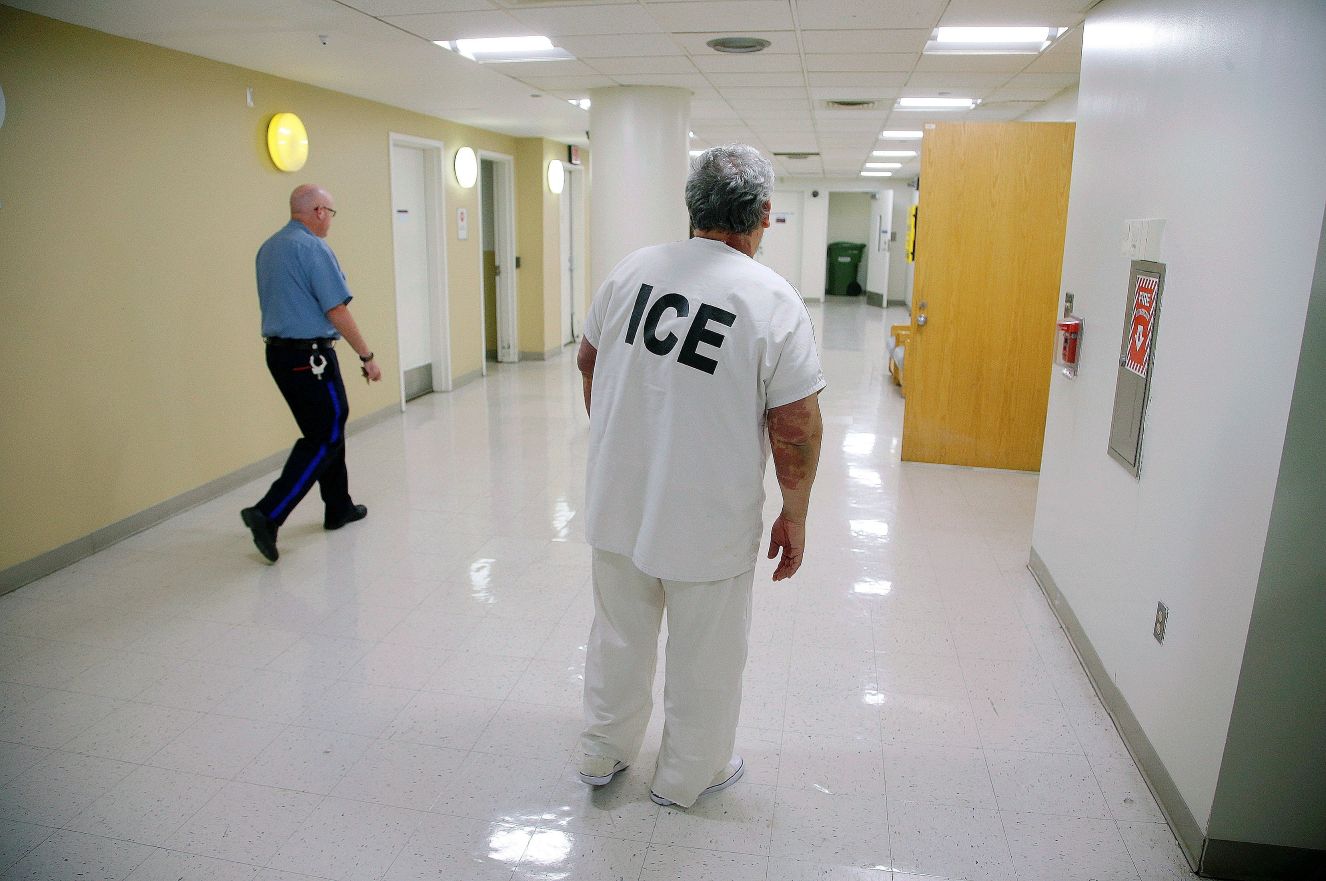 An ICE detainee is escorted by a guard down a hallway.