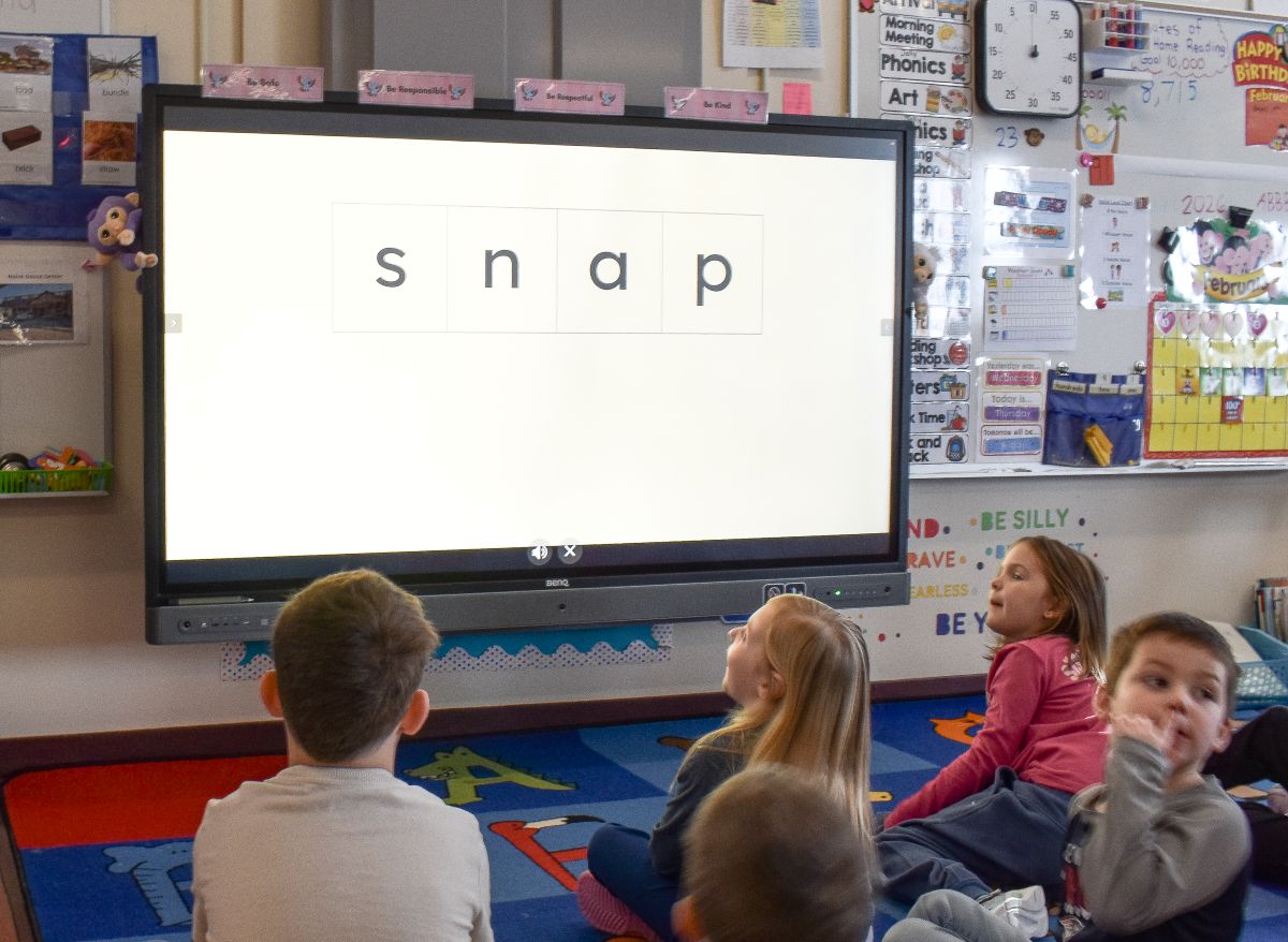 students sit on the floor and look at a large screen that is displaying the word "snap" 