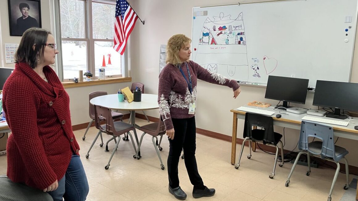 Two women in a classroom space. One is pointing to a desk with computers.