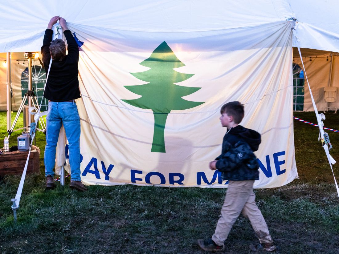 an individual hangs a white Pray for Maine banner that features a green pine tree.