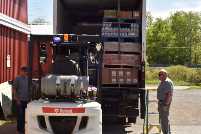Two men watch as a pallet is loaded into a box truck.