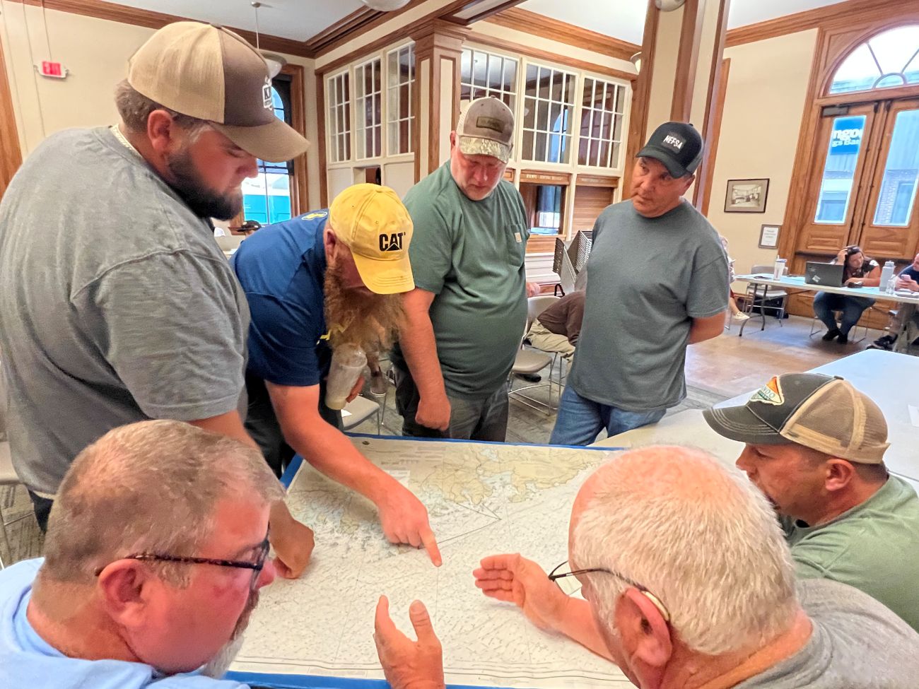 a group of council members stand and sit around a map, with some pointing to various points on the map.
